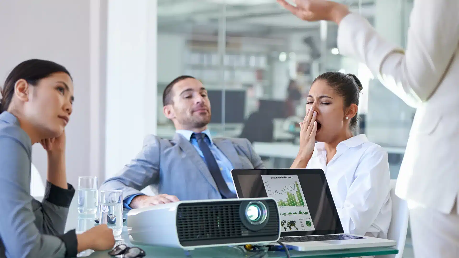 Foto de reunião corporativa: três pessoas demonstram cansaço e desinteresse enquanto alguém apresenta; sobre a mesa há projetor e notebook com gráficos de desempenho.